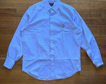 May include: Light blue button-down shirt with long sleeves. The shirt has a button-down collar and a small embroidered emblem on the chest. The shirt is made of a light-colored fabric and is laid flat on a wooden surface.