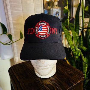 Op de afbeelding: Zwarte baseballcap met de letters "FD NY" in het rood en een rood-wit-blauw embleem. Het embleem toont een Amerikaanse vlag en de woorden "Eer, Moed, Moed" en "9-11-01".