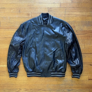 May include: Black bomber jacket with sequined sleeves and a leather-like body. The jacket features a ribbed collar, cuffs, and hem with gray stripes. It has a button-up front and two front pockets. A stylish outerwear piece.