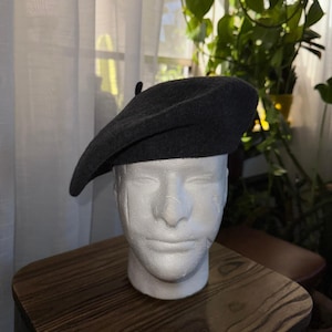 May include: A dark gray beret made of a soft material. The beret is displayed on a white foam head. The beret has a classic, round shape with a small stem on top. The background includes a wooden table and a window with a curtain.