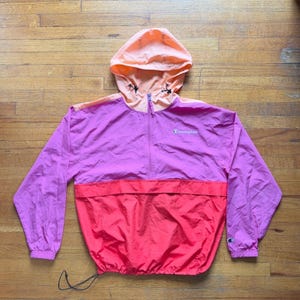 May include: A color-blocked Champion windbreaker jacket featuring a light peach hood with black drawstrings, a magenta upper torso, and a bright red lower section. The jacket has a half-zip front closure, long sleeves with elastic cuffs, and a logo.