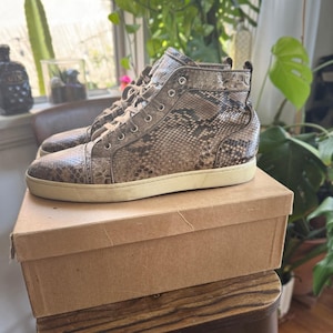 Op de afbeelding: High-top sneakers met een bruin en zwart slangenleerpatroon op het bovenwerk, zilverkleurige metalen oogjes, bijpassende veters en een effen gebroken witte rubberen zool, rustend op een eenvoudige bruine kartonnen schoenendoos.