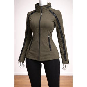 Puede incluir: Chaqueta verde oliva con cuello alto y cremallera completa. Rayas negras con el texto "KITH KITH KITH" recorren las mangas. La chaqueta tiene una silueta entallada y dos bolsillos delanteros con cremalleras negras.