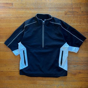 May include: Black and white short-sleeve jacket with a stand-up collar and a quarter-zip closure. The jacket features white piping along the seams and white panels on the sides with zippered pockets. The jacket is made of a lightweight material.