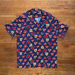 Puede incluir: Una camisa de manga corta con fondo azul y un patrón repetido del logo de Superman en rojo y amarillo. La camisa tiene cuello en V.