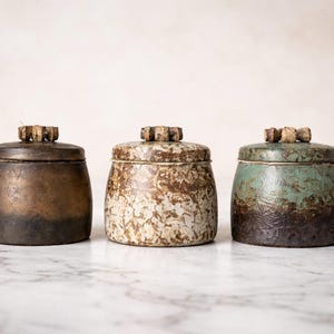 Könnte beinhalten: Drei Keramikgläser mit Deckeln, jedes mit einer einzigartigen Farbe und Textur. Die Gläser sind zylindrisch mit einem dunkelbraunen, gesprenkelten und teal-farbenen Finish. Die Deckel haben einen dekorativen Holzknauf. Die Gläser werden auf einer Marmoroberfläche präsentiert.