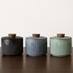 May include: Three ceramic jars with wooden lids. The jars are cylindrical with vertical ribbed detailing. Colours include black, blue-grey, and pale green. The jars are displayed on a dark brown surface against a white background.