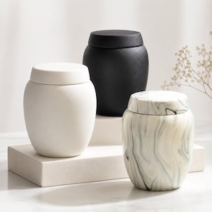 May include: Three ceramic jars with lids, each featuring a distinct finish: one matte white, one matte black, and one with a white and grey marbled pattern. These rounded containers have a smooth texture and are arranged in a tiered display.