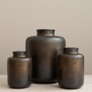 May include: A set of three bronze-effect ceramic vases. The vases have a rounded shape and dark lids. The largest vase is in the centre, with two smaller vases flanking it. The finish has a speckled, metallic appearance, creating a warm aesthetic.