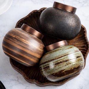 Op de afbeelding: Drie keramische potten met bronzen deksels liggen op een houten dienblad. De potten hebben unieke afwerkingen: één is donker gespikkeld, een andere heeft bruine en bruine strepen, en de derde heeft groene en bruine strepen.