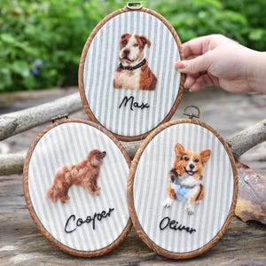 May include: Three oval embroidery hoops featuring needle-felted dog portraits on grey and white striped fabric. Each hoop displays a different dog breed with a name embroidered in black thread below: Max, Cooper, and Oliver.