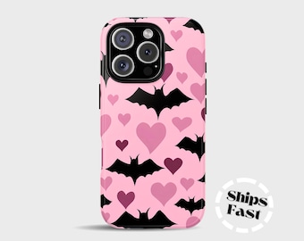 May include: A pink phone case featuring a pattern of black bats and pink hearts. The case has a glossy finish and a black camera module. The words "Ships Fast" are printed on the lower right corner.