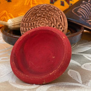 Puede incluir: Un cuenco rojo de terracota está en primer plano, con un objeto circular marrón tallado detrás. También se ven un cuenco pequeño marrón y una pieza decorativa de cerámica marrón. El fondo presenta un textil amarillo con detalles bordados.