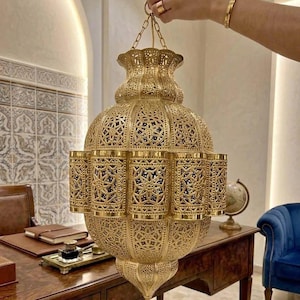 Könnte beinhalten: Eine große, verzierte goldfarbene Laterne mit aufwendigen Ausschnittmustern. Die Laterne, an einer Kette aufgehängt, hat eine bauchige Form mit zylindrischen Seitenakzenten. Das Design deutet auf einen orientalischen oder marokkanischen Stil hin, geeignet für die Inneneinrichtung.