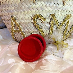 Puede incluir: Un bolso tote de paja tejida con las letras "ASM" en adornos dorados y perlados. Tres peque&ntilde;os recipientes redondos de terciopelo rojo est&aacute;n frente a la bolsa, junto con un peque&ntilde;o regalo envuelto en papel blanco y atado con una cinta dorada.