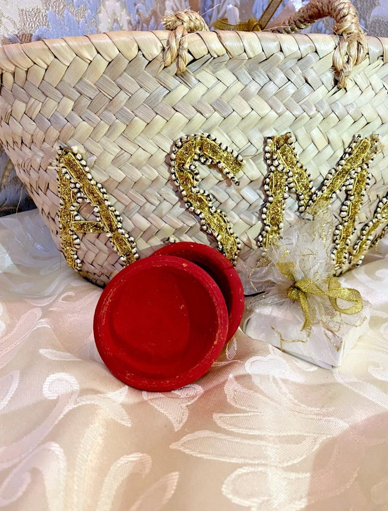 Puede incluir: Un bolso tote de paja tejida con las letras "ASM" en adornos dorados y perlados. Tres peque&ntilde;os recipientes redondos de terciopelo rojo est&aacute;n frente a la bolsa, junto con un peque&ntilde;o regalo envuelto en papel blanco y atado con una cinta dorada.