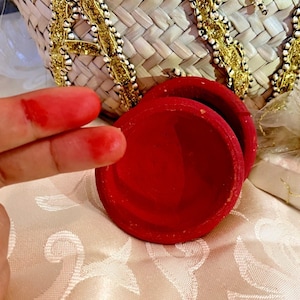 Puede incluir: Dos pequeños recipientes redondos de color rojo brillante, posiblemente para guardar especias o objetos pequeños. Están colocados sobre una superficie color crema, con una cesta tejida y adornos dorados en el fondo.