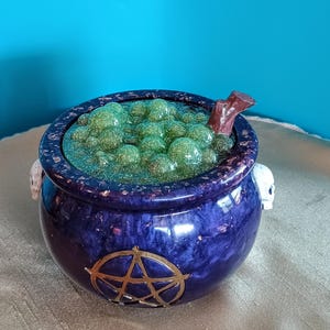 May include: A dark blue cauldron with a gold pentagram design on the front. The cauldron is filled with green, bubbly liquid and has a brown stick and two skull handles. A decorative item for Halloween or a witchy theme.