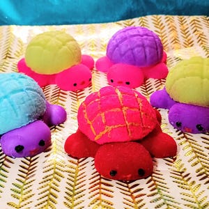 May include: A collection of colorful turtle-shaped bath bombs. The turtles have textured shells in shades of yellow, purple, blue, and red, with matching colored bodies. Each turtle has small black eyes and is displayed on a patterned surface.