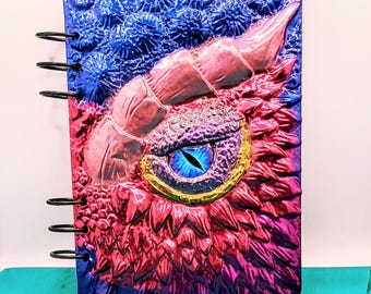 May include: A fantasy-themed notebook with a dragon eye design. The cover features a blue, pink, and purple color scheme with a textured, embossed appearance. The eye is blue with a gold-colored iris. The notebook is bound with black metal rings.