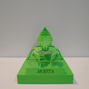 May include: A vibrant green 3D-printed sculpture of an insect-like figure. It is mounted on a rectangular base, with the name "HOJITA" printed on the front. The design includes a leaf-shaped head and multiple legs, all in a vivid green.