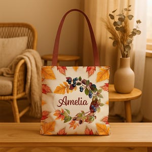 Könnte beinhalten: Beige Tragetasche mit Herbstdesign. Die Tasche hat einen Kranz aus Beeren und Blättern mit dem Namen "Amelia" in der Mitte. Die Griffe sind braun und die Tasche ist mit orangefarbenen, roten und gelben Blättern verziert.