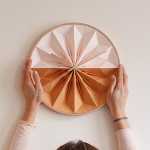 May include: A person is holding up a round wall clock with a pink and orange paper design. The clock has a wooden frame and is made of folded paper.