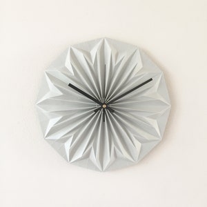 Può includere: Un orologio da parete grigio chiaro, in stile origami, con un design geometrico a forma di stella. L'orologio ha lancette nere e un piccolo centro scuro. L'orologio è montato su una parete bianca, creando un'estetica minimalista.