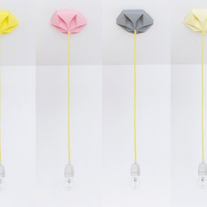 Puede incluir: Cuatro lámparas colgantes de estilo origami en amarillo, rosa, gris y amarillo pálido. Cada lámpara tiene una pantalla del mismo color, un cable amarillo y una bombilla. Las lámparas están montadas sobre un fondo blanco.