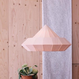 May include: A light pink, geometric, wooden pendant lamp hangs from the ceiling against a light wood background. A small potted plant sits on a wooden table in the foreground.