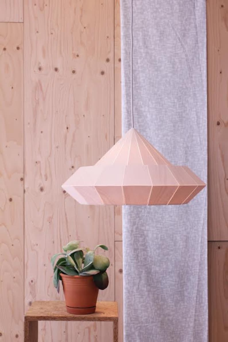 May include: A light pink, geometric, wooden pendant lamp hangs from the ceiling against a light wood background. A small potted plant sits on a wooden table in the foreground.