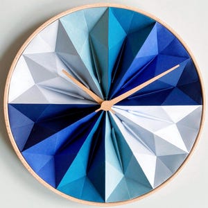 May include: A circular wall clock with a wooden frame and a geometric design. The clock face features folded paper in shades of blue and white, with wooden clock hands. The clock's diameter is approximately 30 cm.
