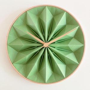Puede incluir: Un reloj de pared redondo con una esfera verde claro de estilo origami y manecillas y marco de madera clara. La esfera del reloj tiene un diseño geométrico de estrella. El reloj es un artículo decorativo para el hogar o la oficina.