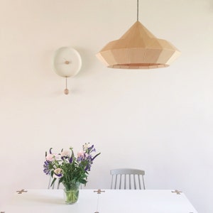 May include: A white dining table with a light wood chair and a bouquet of pink and purple flowers in a glass vase. A white clock with a wooden pendulum hangs on the wall above the table. A large, light wood, geometric pendant light hangs from the ceiling.