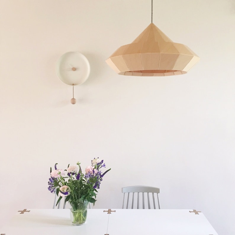 May include: A white dining table with a light wood chair and a bouquet of pink and purple flowers in a glass vase. A white clock with a wooden pendulum hangs on the wall above the table. A large, light wood, geometric pendant light hangs from the ceiling.