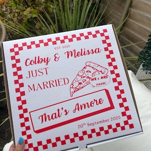 Op de afbeelding: Een vierkante witte pizzadoos met een rood geblokte rand. Het deksel bevat rode tekst met "Colby & Melissa", "EST 2025", "JUST MARRIED", "That's Amore" en "20th September 2025". Een pizzagrafiek bevat de tekst "MR & MRS TIGGES".