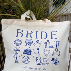 May include: Cream-colored tote bag with the word "BRIDE" in blue. The bag features blue illustrations of a disco ball, dog, hand, wine, bow, bowl, boombox, microphone, sun, purse, Eiffel Tower, record, cats, and a person. Text at the bottom reads "15-17th August Brighton."