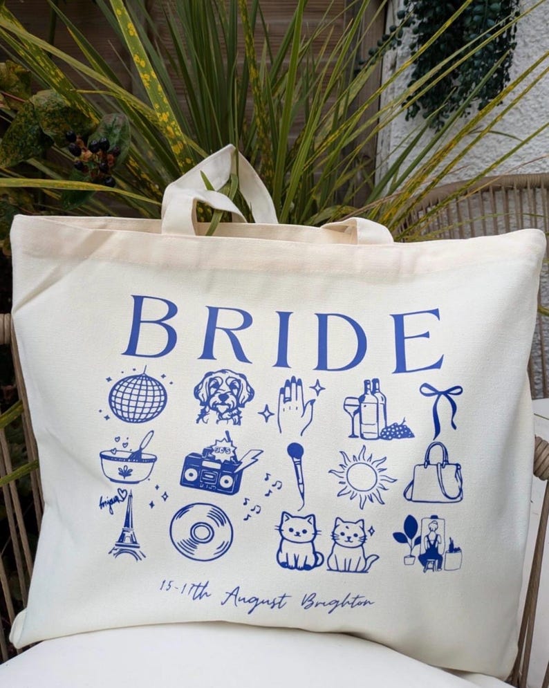 May include: Cream-colored tote bag with the word "BRIDE" in blue. The bag features blue illustrations of a disco ball, dog, hand, wine, bow, bowl, boombox, microphone, sun, purse, Eiffel Tower, record, cats, and a person. Text at the bottom reads "15-17th August Brighton."