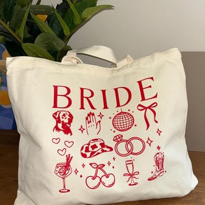 May include: Cream-colored tote bag with the word "BRIDE" in red, along with red illustrations of a dog, hand, disco ball, bow, hearts, cowboy hat, rings, cocktail, cherries, champagne flute, and a boot.