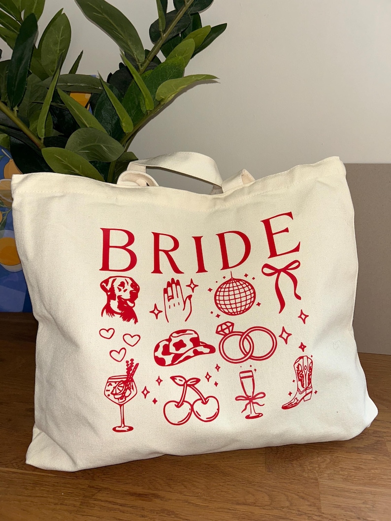 May include: Cream-colored tote bag with the word "BRIDE" in red, along with red illustrations of a dog, hand, disco ball, bow, hearts, cowboy hat, rings, cocktail, cherries, champagne flute, and a boot.