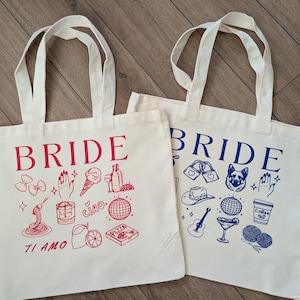May include: Two off-white tote bags with the word "BRIDE" printed in red and blue. Each bag features a collection of hand-drawn illustrations in the same color as the word "BRIDE". One bag has Italian-themed drawings, the other has a variety of drawings.