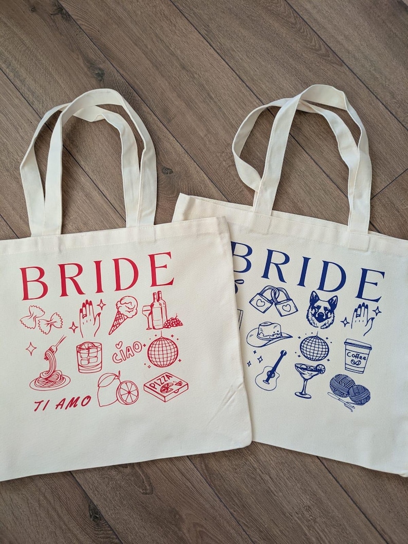 May include: Two off-white tote bags with the word "BRIDE" printed in red and blue. Each bag features a collection of hand-drawn illustrations in the same color as the word "BRIDE". One bag has Italian-themed drawings, the other has a variety of drawings.