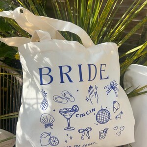 May include: Off-white canvas tote bag with blue text and illustrations. The word "BRIDE" is at the top, with drawings of a pineapple, cocktail, and other beach-themed icons. The text "LAST TOAST ON THE COAST..." is at the bottom.