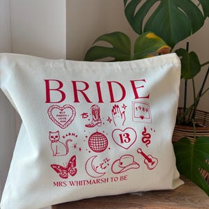 May include: Off-white tote bag with the word "BRIDE" in red. The bag features various red illustrations, including a heart, cat, butterfly, cowboy boot, disco ball, and the text "MRS WHITMARSH TO BE".