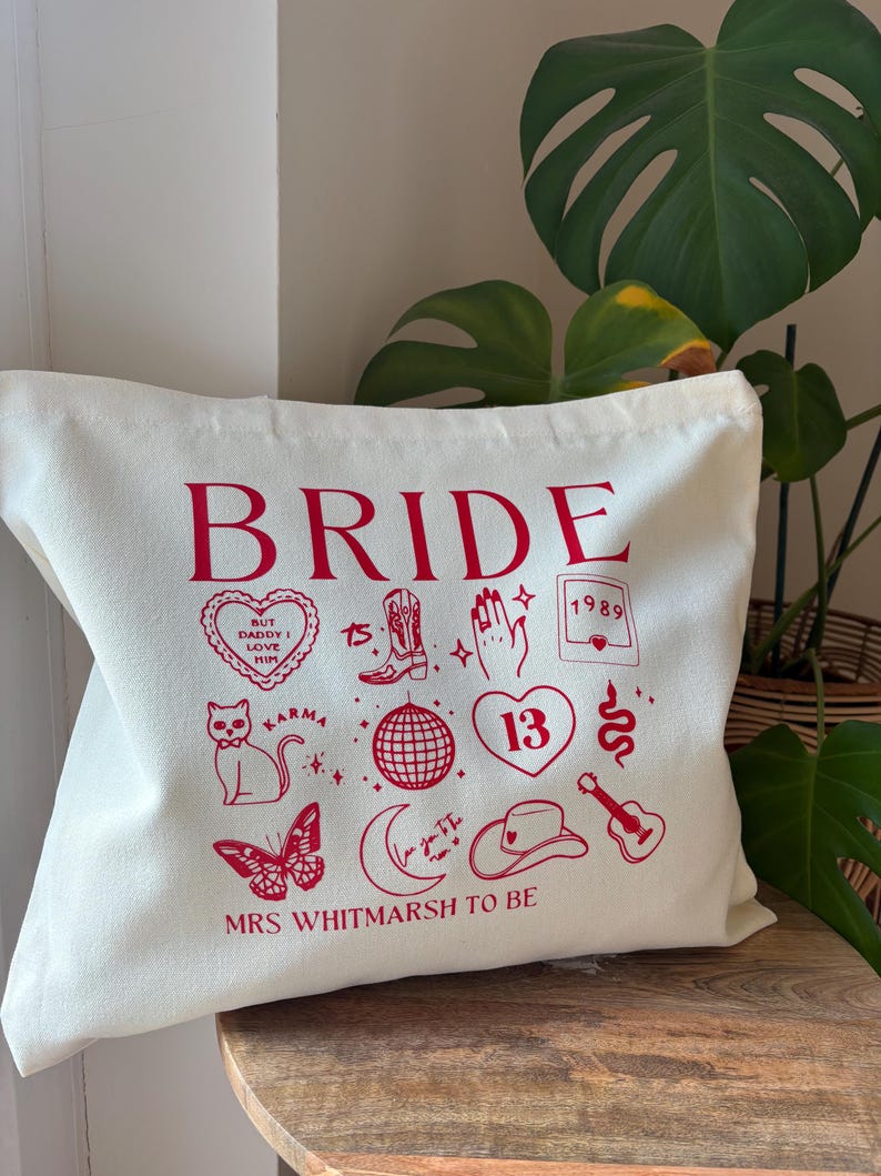 May include: Off-white tote bag with the word "BRIDE" in red. The bag features various red illustrations, including a heart, cat, butterfly, cowboy boot, disco ball, and the text "MRS WHITMARSH TO BE".