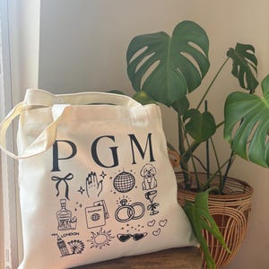May include: Cream-colored tote bag with black illustrations and the letters "PGM". The bag features drawings of a bow, hand, disco ball, dog, cocktail, tequila bottle, passport, rings, London skyline, sun, sunglasses, and hearts. The bag is on a wooden stool.