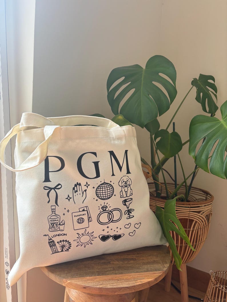 May include: Cream-colored tote bag with black illustrations and the letters "PGM". The bag features drawings of a bow, hand, disco ball, dog, cocktail, tequila bottle, passport, rings, London skyline, sun, sunglasses, and hearts. The bag is on a wooden stool.