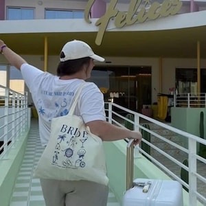May include: A person wearing a white t-shirt and a white cap, carrying a canvas tote bag that says "BRIDE" with various illustrations. The person is also pulling a white suitcase with a gold handle. The background features a building with the words "LOS Felices".