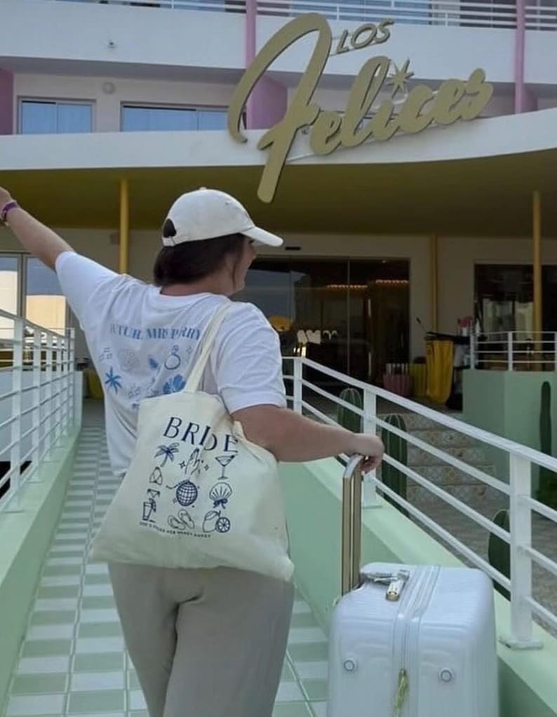 May include: A person wearing a white t-shirt and a white cap, carrying a canvas tote bag that says "BRIDE" with various illustrations. The person is also pulling a white suitcase with a gold handle. The background features a building with the words "LOS Felices".