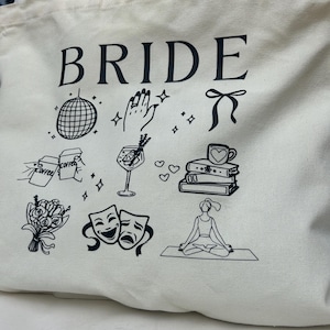 May include: Cream-colored tote bag with the word "BRIDE" in large black letters. The bag features black line drawings of a disco ball, a hand with a ring, a bow, coffee, a cocktail, books, flowers, theater masks, and a person meditating.